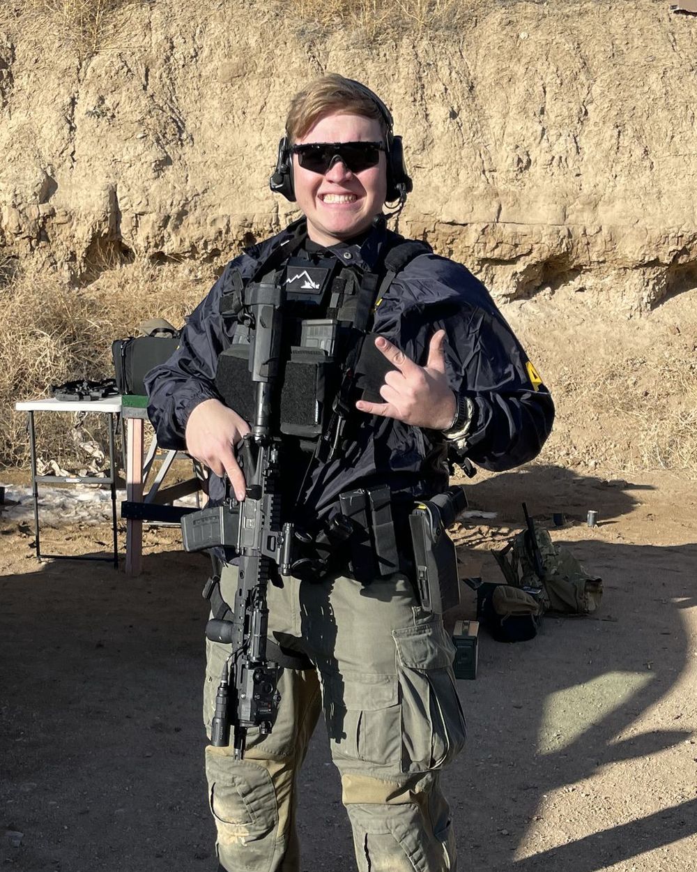 Kyle Milan smiling at an outdoor range in eye and ear protection, with an AR-15 slung muzzle-down