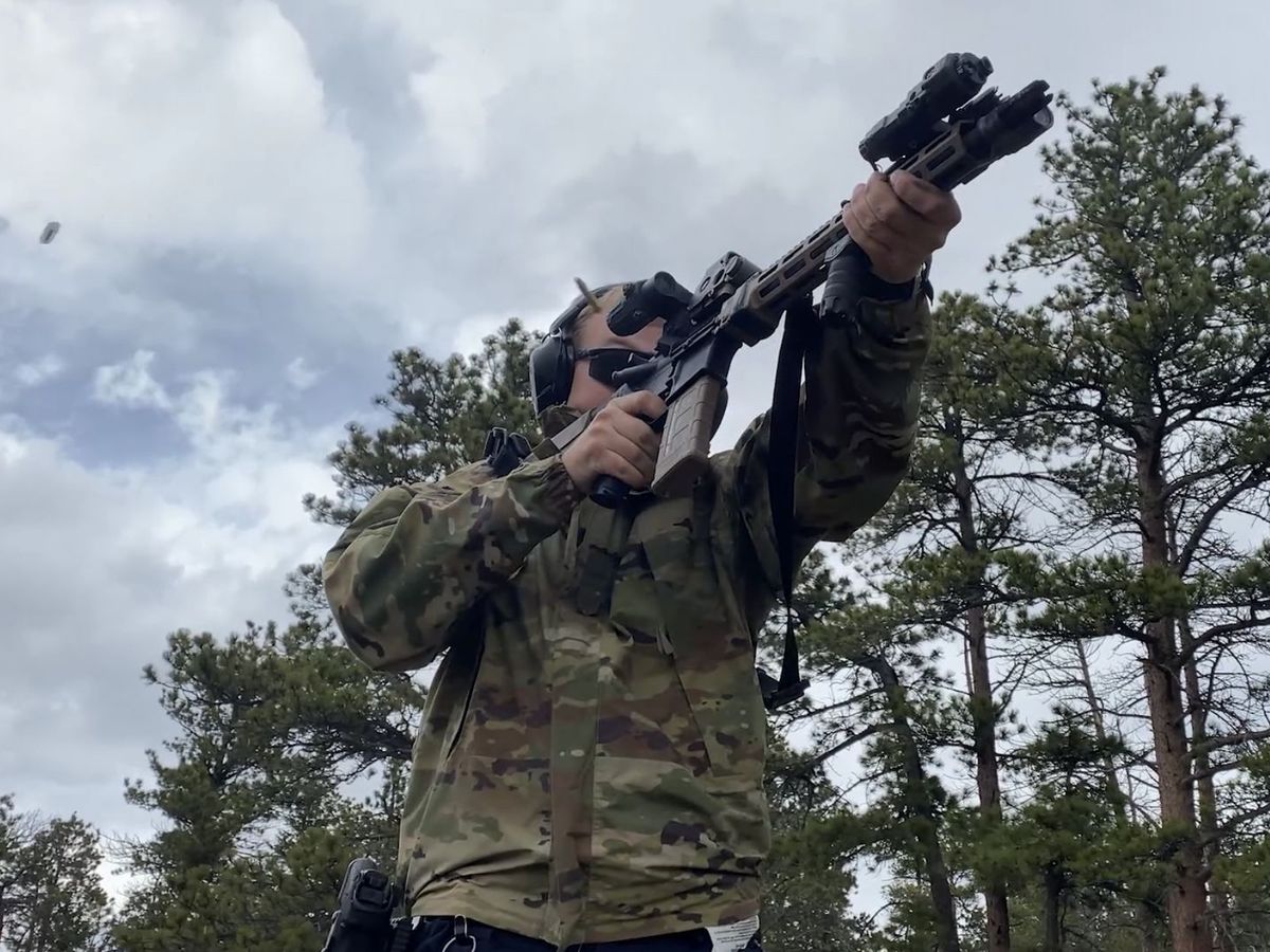 Kyle firing an AR-15 at the range among pine trees, wearing eye and ear protection, with a spent casing ejecting